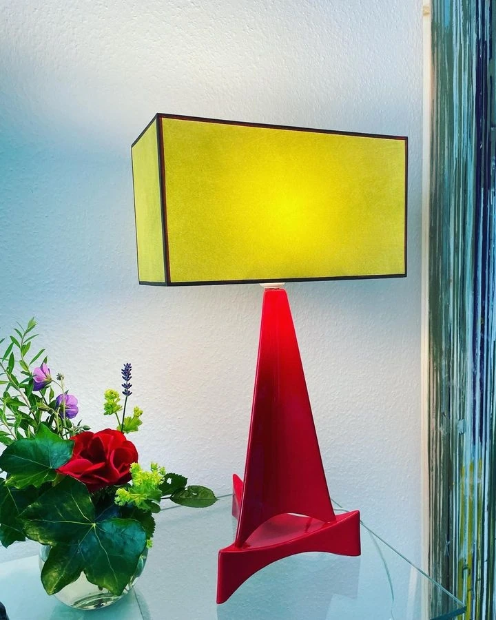 Tischleuchte mit rechteckigem gelben Schirm auf rotem konischem Sockel neben Blumen