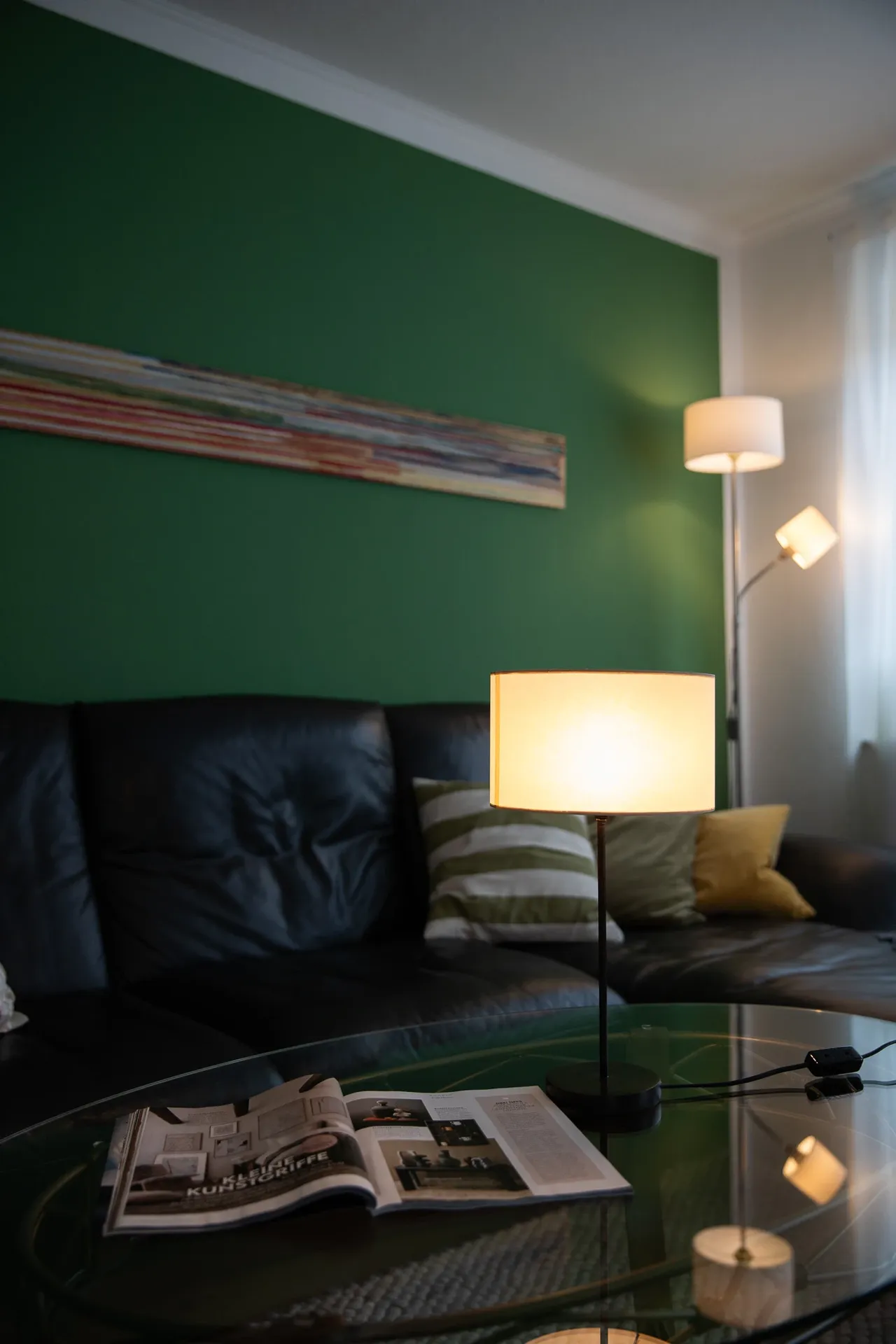 Wohnzimmer mit grün gestrichener Wand, dunklem Ledersofa und beleuchteter Tischlampe