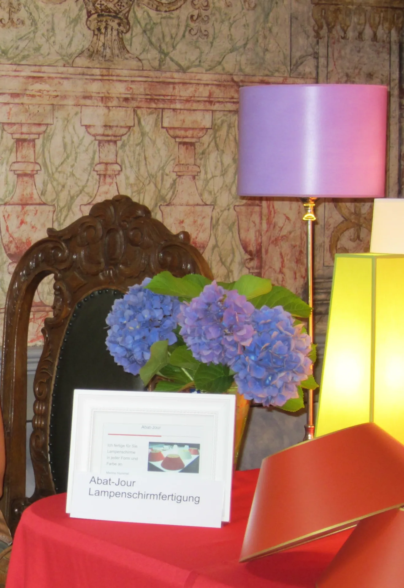 Tischpräsentation mit violettem Lampenschirm, gelber Stehleuchte, Hortensien und Produktkarte