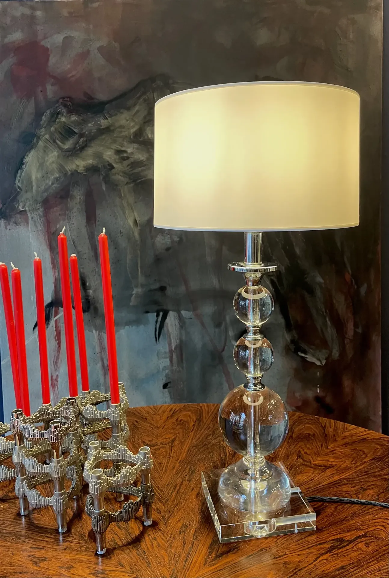 Tischlampe mit klarem Glasfuß und cremefarbenem Schirm neben Metall-Kerzenleuchter mit roten Kerzen