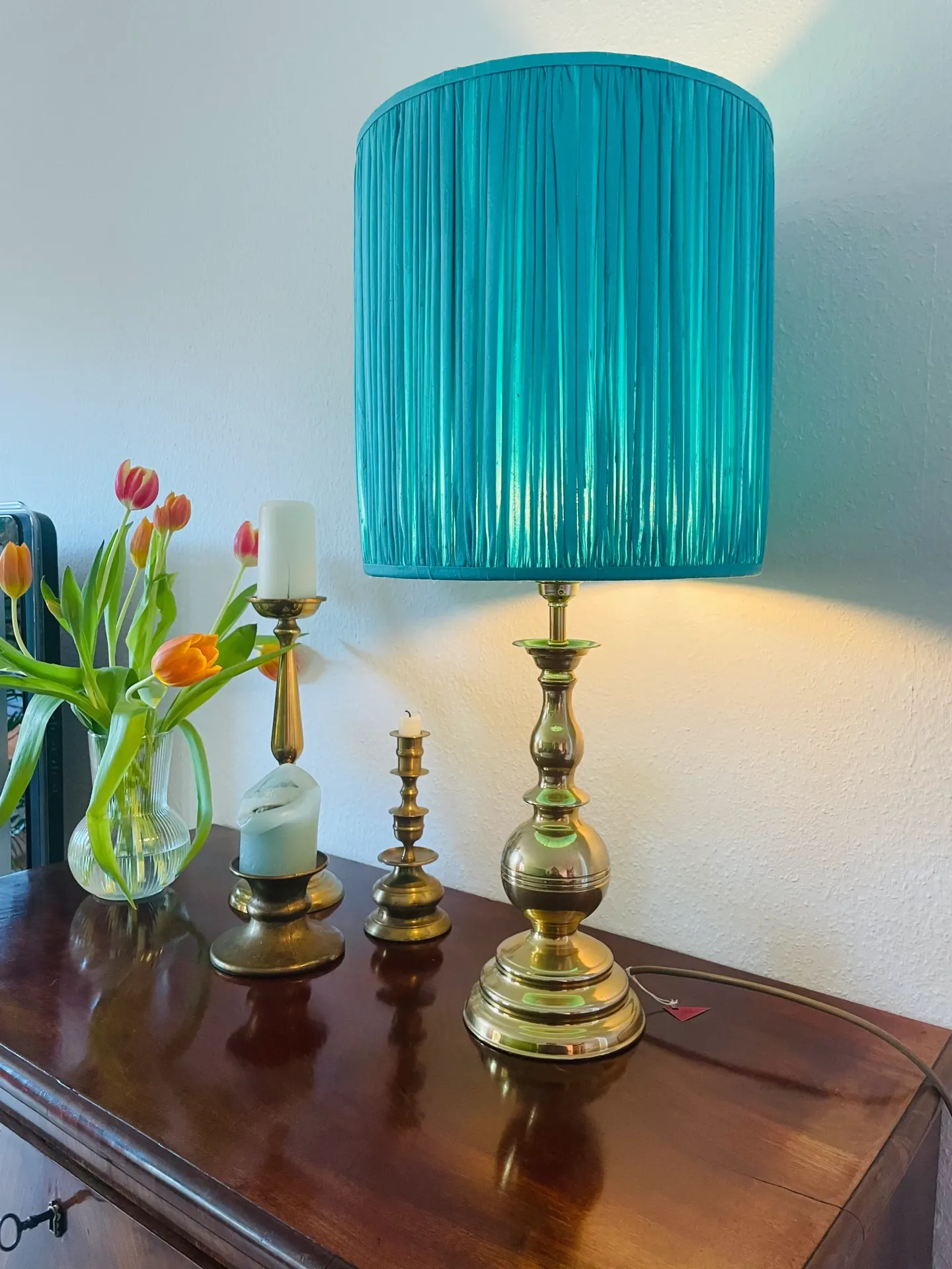 Messinglampe mit türkisfarbenem, plissiertem Schirm auf Holzkommode neben Tulpen und Kerzenhaltern
