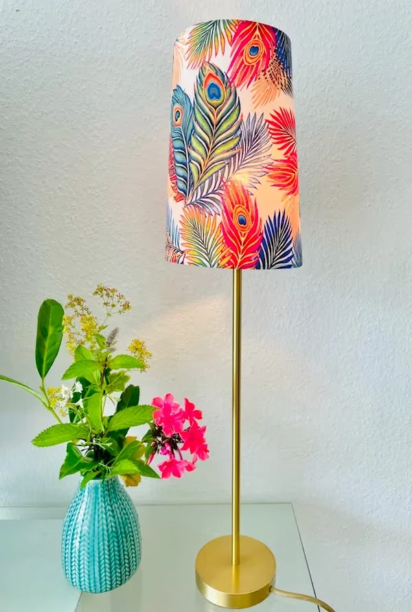 Schlanke Tischlampe mit buntem Pfauenfeder-Schirm neben Vase mit Blumen