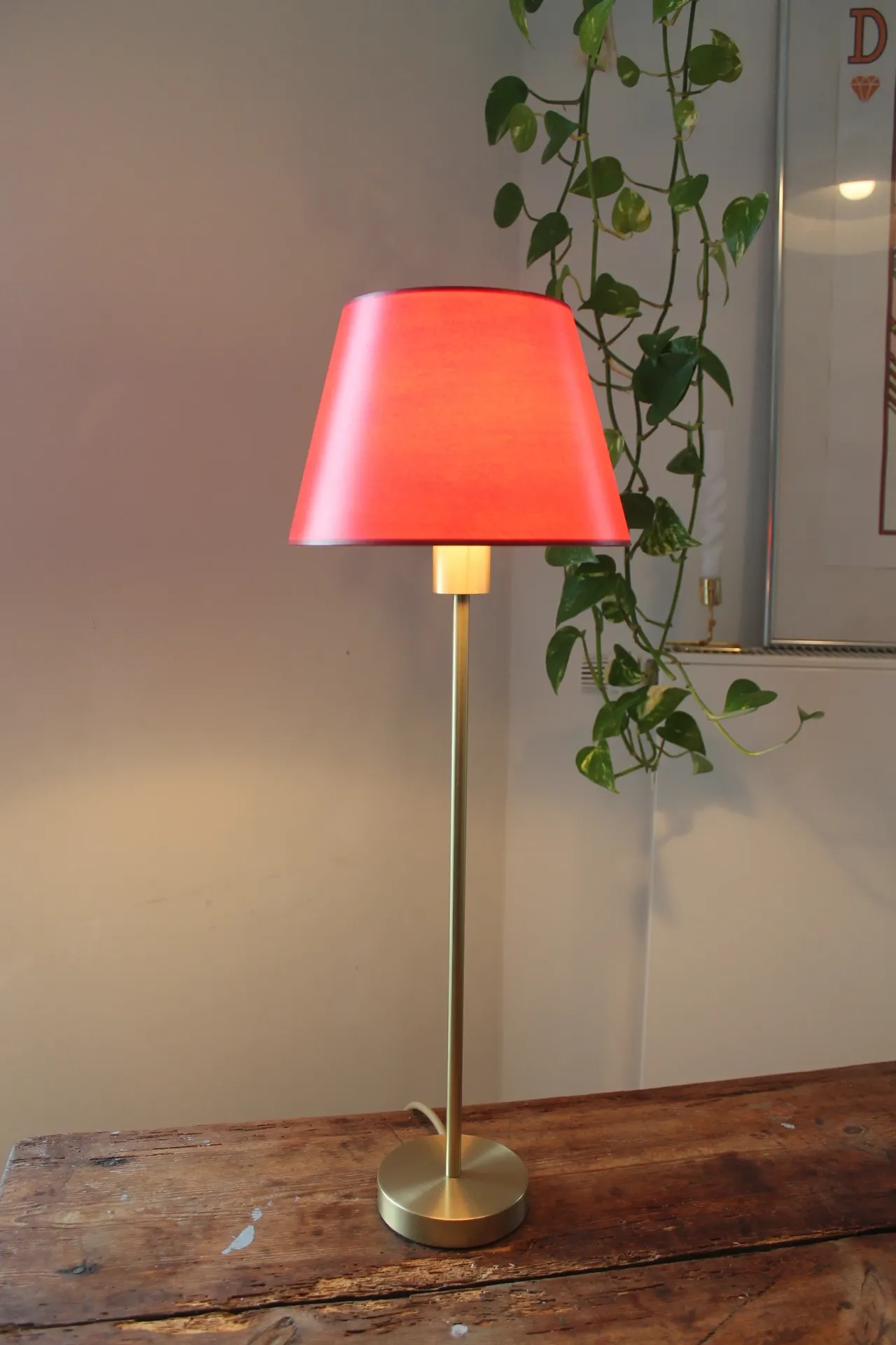 Tischlampe mit rotem Lampenschirm und schlankem Messingfuß auf Holztisch