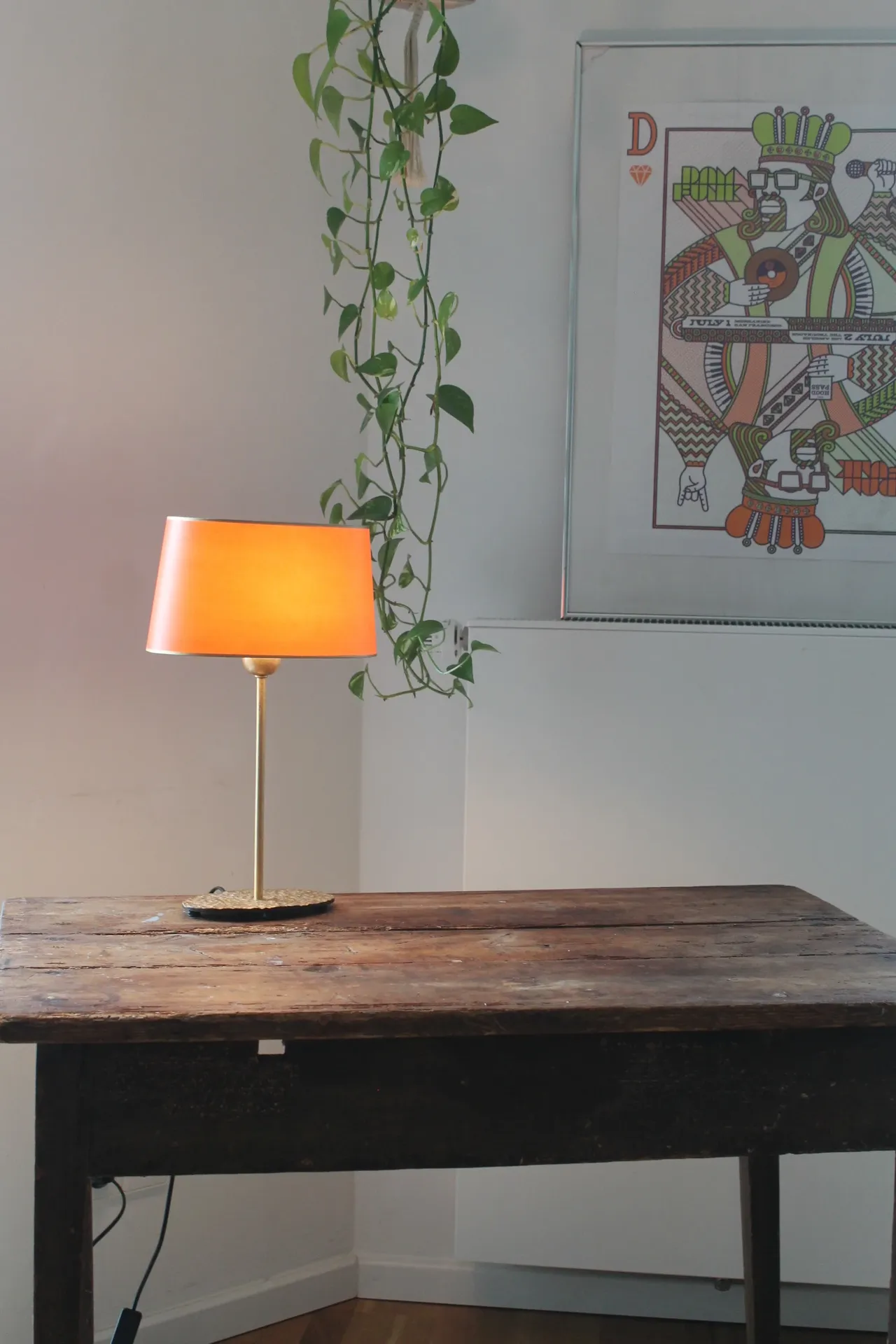 Orangefarbene Tischlampe auf rustikalem Holztisch neben Hängepflanze und gerahmtem Poster