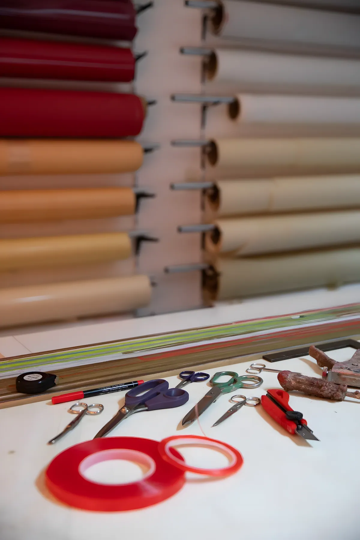 Arbeitsfläche mit Klebebändern, Scheren und Werkzeug vor bunten Rollen an der Wand
