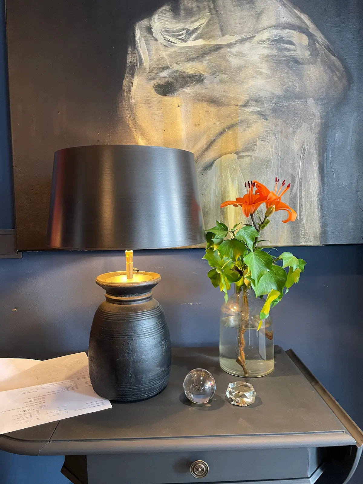 Schwarzer Lampenschirm auf keramischer Lampe auf Tisch, daneben Vase mit orangefarbener Lilie